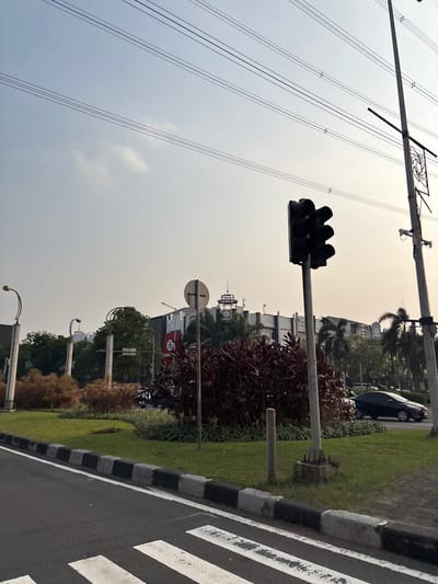 The image depicts an outdoor scene in Pakulonan Barat, Indonesia, featuring a traffic intersection with a pedestrian crossing in the foreground. A traffic light, currently displaying blacked-out signals, stands prominently to the right of the image. Adjacent to the traffic light is a utility pole with intricate metalwork. A black car is visible on the road to the right, moving away from the viewer. The background reveals buildings, including a structure with a red circular logo, and a cluster of palm trees. The sky is hazy and overcast. The area includes a well-maintained grassy median with ornamental plants and trees, bordered by a sidewalk with black and white markings. Overhead, power lines crisscross the sky, supported by poles. No people are visible in the frame.