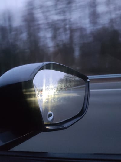 The image depicts a view from a moving vehicle, focused on its black side mirror and the scenery beyond, specifically in Assevillers, France. The side mirror, positioned prominently in the lower-left to mid-right of the frame, is wet with water droplets or streaks, causing the light reflected within to refract and scatter. Bright, white or yellowish headlights from a vehicle behind are clearly visible in the mirror's reflection, creating a strong glow. A small, indistinct white or silver object is also reflected near the bottom center of the mirror.
Beyond the mirror, the background elements are heavily blurred horizontally, conveying a strong sense of motion, typical of driving at speed. This blurred background consists of dark, silhouetted trees, suggesting a forested or rural landscape alongside the road. The sky above is overcast and grey. A metal guardrail runs parallel to the road on the right side of the image, indicating a highway or major thoroughfare.
The lighting conditions suggest twilight—either dawn or dusk—due to the low ambient light and the necessity of vehicle headlights. The presence of water on the mirror hints at recent rain, mist, or high humidity. No people are visible, and there is no discernible text within the image. The overall scene evokes a quiet, somewhat atmospheric moment of travel.