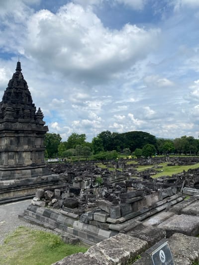 A stone temple, weathered and gray, stands on a grassy plain. The temple is a tall, multi-tiered structure with intricate carvings. The sky is a bright blue with puffy white clouds. The temple is surrounded by a field of rubble and stone blocks, suggesting it is a ruin. The ground is covered in green grass. There are trees in the background. The location is at the coordinates: -7.753 latitude and 110.493 longitude, which is at the Candi Sewu temple complex in Kecamatan Prambanan, Indonesia. The temple appears to be a part of the Candi Sewu temple complex.