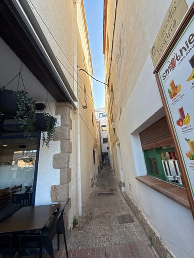 This is a photograph taken in Tossa de Mar, Spain, depicting a narrow, charming alleyway with buildings on either side. The alleyway appears to be a pedestrian walkway, with cobblestone and brick paving. The buildings are made of stucco and stone, with some showing signs of age.
The image is taken from a low angle, looking upwards into the alley. The sky is visible at the top, suggesting it is daytime. The sun is casting shadows, and the light suggests it might be morning or late afternoon. There are no people clearly visible in the scene, but there are hints of human activity.
On the left side of the alley, a sign for "Smoothie By SUCS" is partially visible, advertising various smoothie flavors like "TROPICAL" (pineapple, papaya, mango) and "VITALITY" (strawberry, banana). Below this sign, another sign reads "Passaice Peixeters". This indicates the presence of a food or drink establishment.
Further down the alley, we see an outdoor seating area with dark-colored chairs and tables, suggesting a cafe or restaurant. Hanging plants in dark baskets add a touch of greenery to the scene. The buildings have windows and some architectural details, like decorative stone elements above a white stucco section. Reflections in a large window suggest the presence of shops or interiors on the ground floor.
The overall impression is of a picturesque, slightly weathered European street scene, typical of a historic coastal town. The focus is on the architecture, the signs of local businesses, and the atmosphere of a relaxed, sunny day in Spain.
