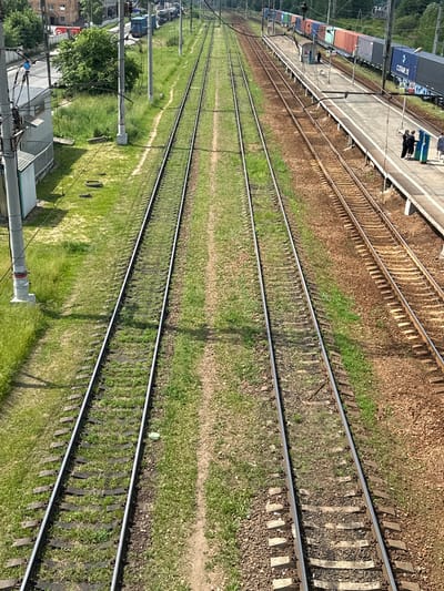 An aerial perspective captures multiple train tracks at a railway station in Selyatino, Russia. The foreground features two sets of parallel train tracks, one on the left flanked by green grass and the other on the right bordered by brown ballast and a concrete platform. In the midground, several cargo train cars, including shipping containers of various colors, are parked on the tracks along the platform. A small blue and gray structure, possibly a signal box or waiting shelter, is situated on the platform. In the background, more train cars and tracks are visible, along with overhead electrical wires and poles, and some buildings and trees lining the railway. Two individuals in dark clothing are standing on the platform, and another person in a blue shirt stands further down the platform. A few more individuals are visible further in the background on the platform. The scene is illuminated by daylight, casting shadows on the tracks and platform.