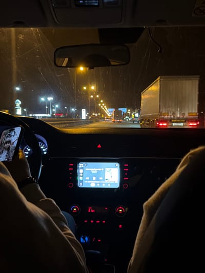 The image captures an interior view from a moving vehicle at night, in Pavlovskaya, Russia, looking forward through the windshield. A person, presumably the driver, has their left hand on the steering wheel and holds a smartphone in their right hand. A black wristband or watch is visible on the left wrist. The smartphone screen displays an image of an individual. The vehicle's central infotainment screen is illuminated, showing a music player interface with the title "Памятный романс" and album art. Below it, the climate control displays "21.0C," and the red triangular hazard light button is illuminated. Outside the vehicle, the road is brightly lit by numerous yellow streetlights extending into the distance. A large white semi-trailer truck with illuminated red taillights is visible in the adjacent lane to the right. Additional vehicle lights are present further down the road, and a lit commercial establishment, possibly a gas station, is visible on the left. A partial view of a passenger's leg, clad in light-colored fabric, is present on the far right of the frame. The windshield exhibits streaks or marks.