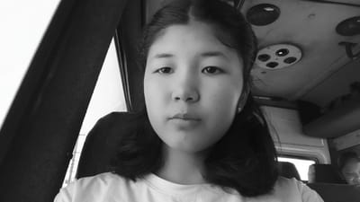 A black and white, medium shot, selfie-style photograph shows a young East Asian female with dark hair styled in pigtails. She is wearing a white t-shirt and a small earring in her right ear. The background is the interior of a vehicle, possibly a bus, with seats visible. A circular vent with four openings is present on the ceiling. In the background, another person is visible in a seat behind the main subject. The lighting is bright, suggesting it is daytime. The image does not provide any specific location information within Kaarman, Kyrgyzstan.