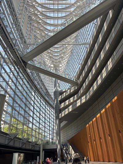 The image captures the interior of the Tokyo International Forum in Chiyoda, Japan, a modern architectural marvel characterized by its vast glass atrium and complex, interwoven steel structure. The perspective is from a ground-level view looking upwards and diagonally across the expansive space.

Dominating the scene is the soaring glass facade on the left, composed of a grid of rectangular panes reflecting the exterior sky and surrounding buildings. Sunlight streams through the glass and the intricate, skeletal ceiling structure, casting geometric shadows and highlights across the interior. The ceiling itself is a symphony of curved steel beams and translucent panels, creating a sense of lightness and openness.

On the right side of the atrium, a multi-tiered structure rises, clad in light-colored textured panels. Diagonal wooden slats form a striking, sloped wall on the lower right, bathed in warm sunlight and creating a strong visual contrast with the metallic and glass elements.

Several escalators are visible, carrying people between levels, hinting at the building's function as a public venue. A large, central pillar with a grey, cylindrical base supports the overhead structures.

In the lower portion of the image, several individuals are present, some walking, others on escalators. Their presence adds a sense of scale to the immense architecture. A group of people near the bottom center, some with brightly colored hair, suggests a public event or gathering. The overall atmosphere is one of grand, contemporary design, flooded with natural light.