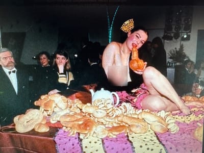 In the city of Protasovo, Russia, a woman is posed with a phallic-shaped object, her mouth open and touching it. She is wearing a bright yellow and black polka dot bow in her hair. She is partially undressed, with her torso exposed. She is sitting on a bed or table covered in a colorful, polka-dotted fabric. In front of her are numerous bread rolls, some of which are also phallic-shaped. To her left, a man in a tuxedo and bow tie looks on, his expression neutral. Other people are visible in the background, mostly out of focus, suggesting a crowded event or party. A white teapot sits amidst the bread rolls. The overall scene appears to be a staged, provocative display.