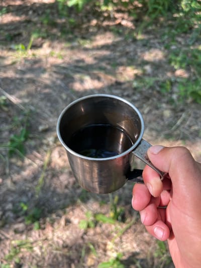 A close-up shot shows a hand holding a stainless steel mug filled with a dark liquid, likely coffee or tea. The mug has a simple, utilitarian design with a small attached handle. The hand, with visible fingernails, is grasping the handle and the side of the mug. The background is a blurred outdoor scene with dappled sunlight filtering through what appears to be trees, creating shadows on the ground. Patches of green foliage are also visible in the soft-focus background. The overall impression is of a moment of quiet enjoyment outdoors. The location in Novohnide, Ukraine is not discernible from the image.