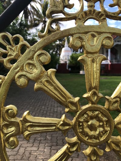 The image captures a close-up perspective of an ornate, golden metal gate or railing component in the foreground, partially obscuring the background. The metalwork is intricately designed with a central circular medallion featuring a stylized floral or rosette pattern, surrounded by eight radial spokes that terminate in various decorative elements, including scrolls, pointed tips, and bulbous forms. The surface of the golden metal appears textured, possibly cast iron with a hammered or painted finish. A black horizontal bar, likely part of the gate frame, is visible at the top left.

In the mid-ground and background, the scene reveals a path made of interlocking grey paver blocks leading towards a grassy area. Beyond the grass, a prominent white stupa or dagoba with a tiered, pointed finial is visible to the left. To the right of the stupa, part of a building with a reddish-brown tiled roof and white walls can be seen. The background features lush green foliage, including palm trees with their fronds silhouetted against a bright, partly cloudy blue sky. No individuals are present in the scene. The overall environment suggests a temple or monastery ground in Karandugoda, Sri Lanka.