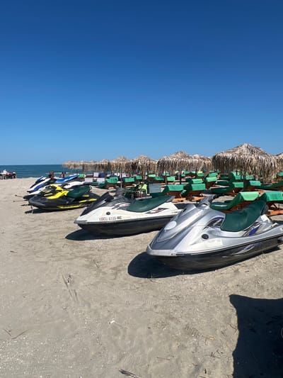 The image shows a row of jet skis parked on a sandy beach, with the blue ocean and sky in the background. Several thatched umbrellas are also visible, providing shade over beach chairs. The scene suggests a sunny day, likely during the warmer months, with the jet skis ready for recreational use. Some of the jet skis are white and silver, while others are black and yellow. One jet ski has a license plate that reads "ROU-0725-MC". The sand appears to be a mix of fine grains and small shells. In the distance, the sea stretches out towards the horizon. A shadow of a person can be seen on the sand. The overall impression is one of a typical beach resort scene, with water sports available for visitors. The location is identified as Năvodari, Romania, which is a coastal town known for its beaches.