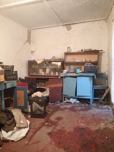 The image depicts an interior scene, likely a room in a dwelling, in Veremia, Ukraine. The environment appears neglected and shows signs of disrepair. The walls are a light color, possibly white or off-white, and exhibit significant cracking, particularly in the upper left corner. A portion of the ceiling is visible, also showing signs of wear and staining.
The room is furnished with several pieces of furniture. A large, dark-colored cabinet with glass doors is positioned against the back wall, containing an assortment of glassware and some dishes. Above this cabinet, a wooden shelf unit holds more glassware, dishes, and a jar. In front of the cabinet is a light blue, wooden piece of furniture, possibly a small desk or cabinet, with its doors ajar. To the left of this blue furniture, a smaller, darker blue wooden desk or table is visible, with stacked black plastic crates on top. To the right of the blue furniture, a simple blue wooden chair or bench is situated.
Various objects are scattered around the room, contributing to the appearance of disarray. Black plastic crates are present in multiple locations. A dark-colored bag or bundle of clothing is on the floor in the lower left corner, next to a crumpled white sheet or tarp. The floor itself is a dark, reddish-brown color and appears stained or dirty, with scattered debris and what might be dried liquid stains. In the lower right corner, a broom is leaning against the wall.
The overall impression is one of abandonment or severe neglect, suggesting a dwelling that has fallen into disuse or is in a state of disrepair. There are no visible individuals present in the image.