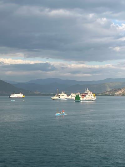 In Upt Tambak Sari, Indonesia, a serene ocean scene unfolds under a dramatic sky. Several large ferries, their white hulls stark against the deep blue water, are anchored or moving slowly. One ferry on the left displays a yellow stripe on its stern and is accompanied by smaller, blue boats. Another, larger ferry in the center, features a green section on its side and is heading towards the right. Further to the right, a third ferry with red and white accents is visible. In the foreground, a small blue boat with a few individuals on board is noticeable. The background is dominated by rolling hills and mountains, their forms softened by the hazy atmosphere and the overcast sky. The water exhibits subtle ripples, reflecting the muted light of the day.