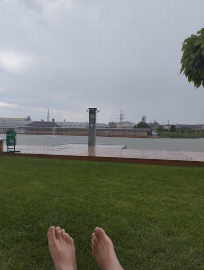This media file shows a ground-level view of a park with a focus on a person's bare feet resting on lush green grass. In the background, a large body of water, possibly a lake or reservoir, is visible, with a wooden pier extending into it. A shower fixture stands on the pier, suggesting it might be an area for swimming or water activities. Beyond the water, a row of buildings with glass facades and covered seating areas can be seen, along with industrial structures and power lines in the distance, indicating an urban or industrial edge to the park. The sky is overcast and grey, and there are visible raindrops on the water and on the wooden pier, suggesting it is raining. The overall atmosphere is subdued due to the weather. No visible text is present in the image. The location context is Romanivka, Ukraine.