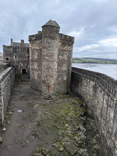The image displays Blackness Castle in Blackness, United Kingdom, under an overcast sky. The primary subject is the castle's robust stone architecture, viewed from an elevated position. The central feature is a prominent cylindrical stone tower, known as the Fore Tower or Stem, with a conical dark grey roof and battlements at its top. This tower features multiple small, rectangular windows with white frames. A section of reddish-brown stone is visible on the lower right side of the tower. Behind and to the left of this tower, another section of the castle, referred to as the Stern, is visible, featuring a dark sloping roof and at least two chimneys.

Two substantial stone curtain walls enclose the castle structures. The left wall has a walkway topped with a dark metal railing. The right wall also features a similar walkway and railing, bordering a wide expanse of dark water, likely the Firth of Forth, which appears to be at low tide, revealing muddy or rocky banks on the distant shore. The ground level within the castle walls, between the central structures and the outer walls, is exposed, showing a muddy, uneven terrain interspersed with rocks and patches of green moss or algae. Several small puddles are present on this ground.

In the background, beyond the water, a gently sloping landscape of green fields and distant trees is visible. The sky is uniformly grey and heavily clouded. Two small, indistinct figures are faintly visible near an arched entrance on the lower section of the main castle building to the left of the central tower; their specific actions or interactions cannot be discerned due to distance and low resolution.