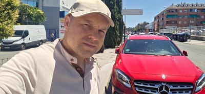 A man wearing a light-colored cap and a beige polo shirt is taking a selfie outdoors. He is positioned on the left side of the frame, with a red Mercedes-Benz car occupying the right side. Behind the man, a white Iveco van is visible. The scene appears to be in a city, with buildings and a street lined with trees and cars in the background. The location is identified as Collado Villalba, Spain. The man's expression is neutral, and he is looking towards the camera. The sun is shining, indicating a clear day. The environment suggests an urban setting, possibly a parking lot or a street with commercial establishments. There are also several large trash bins in the background, along with a parking sign indicating public parking. The overall scene is a snapshot of a person in an urban environment.