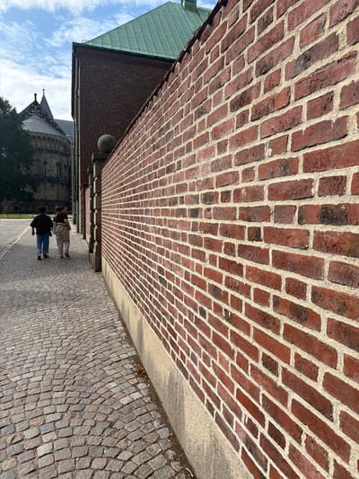 This is a photograph taken at Lund Cathedral in Lund, Sweden. The image shows a cobblestone pathway leading away from the viewer, bordered on the right by a tall, red brick wall. The wall is detailed with visible mortar lines between the bricks. In the distance, on the left side of the pathway, the distinctive architectural features of Lund Cathedral are visible, including its spires and stone facade. Two individuals, appearing to be young women, are walking away from the camera on the cobblestone path, their backs to the viewer. One is wearing a dark top and blue jeans, and the other is wearing a light-colored top and pants. The sky above is blue with scattered clouds. The scene is captured in daylight, suggesting an outdoor setting.