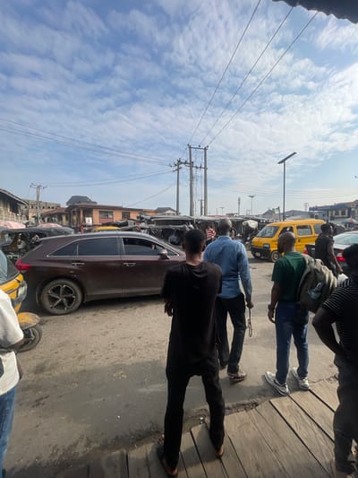 The image depicts a bustling street scene in Akowonjo, Nigeria, under a partly cloudy sky. Several individuals are present, some standing and observing the traffic, while others appear to be walking or waiting. In the foreground, a group of men are seen from behind, some with bags on their shoulders. A brown SUV is parked on the left side of the road, and a yellow van is visible further down the street. The road is paved and appears to be busy with vehicular and pedestrian activity. Buildings line the street, suggesting a commercial or residential area. Power lines crisscross the sky, and streetlights are mounted on poles. The overall environment suggests a typical urban streetscape in Nigeria, characterized by the presence of various modes of transport and the dynamic interplay of people and vehicles.