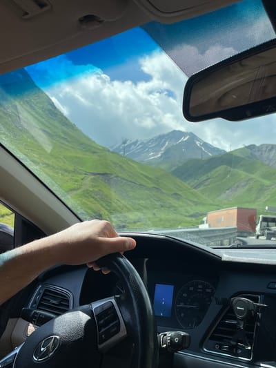 A first-person perspective shows a person's hand on the steering wheel of a car. The dashboard of the car is visible, including a speedometer and a phone mount. Outside the car, a scenic view of lush green mountains is seen through the windshield, with some snow-capped peaks in the distance. A highway with other vehicles, including trucks and cars, is also visible. The sky is blue with scattered white clouds. The scene appears to be set in a mountainous region, possibly on a road trip. The specific location of Almasiani, Georgia, is not discernible from the image itself.
