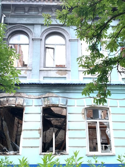 The image shows the facade of a building located at latitude: 49.192, longitude: 37.279 in Izium, Ukraine. The building is light blue with white trim and has five windows. The windows are all broken. The top two windows have their glass panes shattered, while the bottom three windows have been completely blown out. The interior of the building is visible through the broken windows, and there is debris and charred wood inside. The building appears to have been damaged by fire. A tree with green leaves partially obscures the building, but it is growing outside of the structure. The building is likely residential. No people are present.