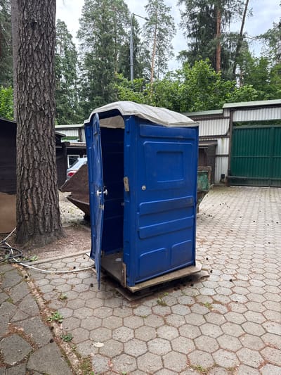 In Сосны, Russia, a blue portable toilet with an open door stands on a hexagonal paver surface. The toilet is positioned next to a large tree trunk with rough bark and to the left of a dark wooden structure. Behind the toilet, a large green metal gate is visible, along with a corrugated metal wall. Further back, tall pine trees form a dense forest. The ground is scattered with pinecones. A silver car is partially visible behind the portable toilet, along with a large, rusty metal container. A light-colored hose is coiled on the ground to the left of the tree. The sky is overcast.