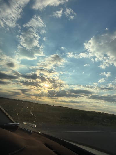 The image captures a scenic view from inside a vehicle, likely a car, looking out at a landscape and sky. The primary subjects are the expansive sky filled with a variety of clouds, and the terrain visible beyond the windshield. Sunlight is breaking through the clouds, creating a dramatic effect with visible sunbeams. The time of day appears to be late afternoon or early evening, given the angle and quality of the light.
The setting is a rural or semi-rural area, suggested by the green fields and vegetation bordering a road. The road itself is visible in the right portion of the frame, with a white line marking its edge. The interior of the vehicle is partially visible, showing a dark-colored dashboard and what appears to be a reflection or some detail on the windshield. The overall mood is serene and awe-inspiring, due to the beautiful interplay of light and clouds. There is no discernible activity or event taking place, other than the passive observation of the external scene from within the vehicle.
Notable details include the textured and varied cloud formations, ranging from wispy cirrus to denser cumulus, illuminated by the setting sun. The sun's rays are a prominent feature, suggesting a moment of atmospheric beauty. The location context is specified as Utrennyaya Dolina, Russia, which implies a Russian landscape, though specific identifying features are not clearly visible. No text is present in the image.