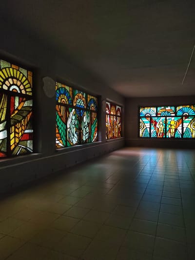 The image shows a long, empty room with tiled flooring. Sunlight streams through large stained-glass windows on the left and right walls, casting colorful patterns on the floor and walls. The windows are adorned with intricate geometric and figurative designs in vibrant hues of yellow, orange, green, blue, and red.

On the left wall, there are three stained-glass panels. The first panel features abstract geometric shapes, predominantly in yellow and orange. The second panel depicts two figures with what appear to be wings, rendered in green and white. The third panel on the same wall also shows figurative elements, possibly people or mythical beings, in warm tones of yellow and orange.

The right wall has a larger stained-glass window with a central design featuring two standing figures in yellow and red, surrounded by fan-like patterns and circular motifs in blue and green. The overall impression is one of solemnity and artistic beauty, likely within a public building or a place of worship, given the nature of the stained glass. The light suggests it is daytime, possibly late afternoon given the long shadows. The location context provided is Свірскі сельскі Савет, Belarus, hinting at a rural or small-town setting within Belarus. There is no visible text in the image.
