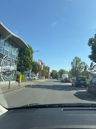 The image shows a street in Ahlen, Germany, with several cars parked along the right side. On the left, there is a building with a large "TREFFPUNKT" logo. The sky is clear and blue, and the trees are green, indicating it is likely daytime during a warm season. There are flags on poles in front of a building further down the street. A white van is parked behind a red car in the middle of the road. A dark blue car is parked to the right of the red car, with a license plate visible. The image is taken from inside a vehicle, looking forward.