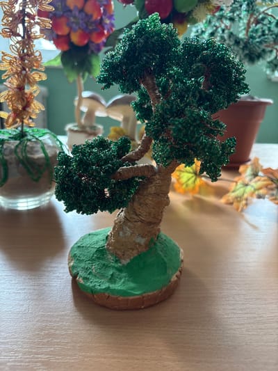 The image shows a close-up of a handmade beaded tree figurine. The tree has a textured, bark-like trunk wrapped in brown and gold material, and its canopy is densely covered with small, shiny green beads. The tree is rooted in a base made of what appears to be clay or a similar material, painted green to resemble grass, and it sits on a light brown wooden surface. In the background, out of focus, are other decorative items, including what looks like another beaded plant or flower with orange and purple elements, and a pair of ceramic mushrooms. A brown planter pot is also visible in the background. The lighting suggests an indoor setting, likely a table. The location is specified as Ветрынскі сельскі Савет, Belarus.