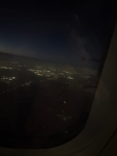The image shows a view from an airplane window at night. Outside, there are faint lights that suggest a city below, potentially Tremblay-en-France, France. The interior of the plane is dark, with the window frame visible. No people or specific objects can be identified in the darkness, and the nature of any actions or interactions is unknown. The environment is limited to the view outside the plane window at night.