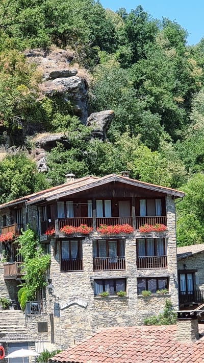 The image shows a stone building with a brown tiled roof, situated on a hillside in Rupit i Pruit, Spain. The building has multiple levels, each featuring balconies with wooden railings and red flowers in planters. Windows with white curtains are visible on each level. A lush green forest with a rocky outcrop serves as the backdrop. The sky is clear and blue. There are no people visible in the image.