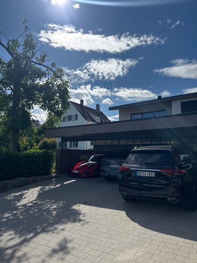 The image shows a modern carport attached to a multi-story white house with a dark tiled roof, located in Brackenheim, Germany. Three cars are parked under the carport. On the far left is a red Porsche sports car with white racing stripes on the hood and roof, bearing the license plate "HN GT 389". To its right is a dark gray BMW SUV with the license plate "HN JL 656E". The third car, on the right, is a black Mercedes-Benz SUV, also with the license plate "HN AA 288E". The scene is bathed in bright sunlight, casting dappled shadows from a large tree onto the paved driveway. The sky is blue with scattered white clouds, and the sun is high in the sky, creating lens flare in the upper left quadrant of the image. Lush green bushes and a stone wall are visible to the left of the carport.