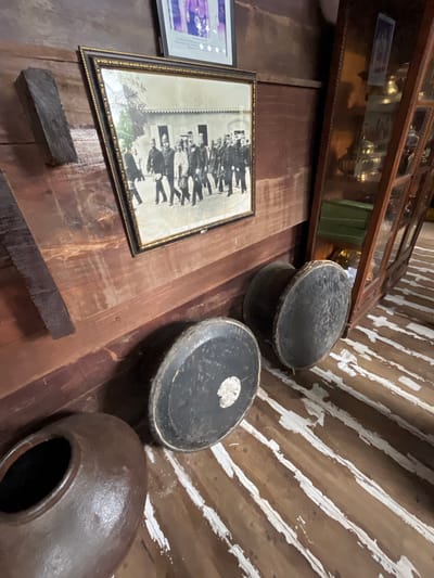 In the city of Mesjidpirak, Indonesia, a dimly lit room features wooden walls and a floor of distressed wooden planks. Dominating the left side of the frame is a framed black and white photograph of a group of men in uniform walking outdoors. To the right of the photograph, leaning against the wall, are two dark, circular, shallow drums, possibly gongs. In the foreground on the left, a large, dark brown, spherical pottery vessel with a wide opening sits on the floor. Further to the right, a tall wooden cabinet with glass doors displays various items, including what appear to be metallic vases and green boxes.