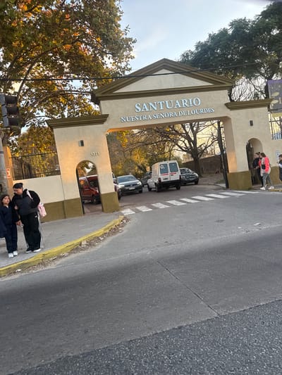The image shows the entrance gate to Santuario Nuestra Señora de Lourdes in Quilmes Oeste, Argentina. The gate is beige with the name of the sanctuary inscribed on it. A street runs past the gate, with a yellow curb and zebra crossing visible. Several parked cars are visible behind the gate. Two people are standing on the sidewalk, near the curb. Other people can be seen standing and talking under the entrance gate. The sky is overcast, and trees are on either side of the gate.