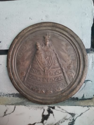 The image shows a circular bronze medallion depicting a religious scene, likely the Virgin Mary and the Child Jesus, set against a textured white wall. The medallion features intricate relief carvings of the figures, adorned with crowns and elaborate clothing. Rays emanate from behind the figures, suggesting a divine aura. The medallion is positioned on a surface with a marble-like pattern, and the surrounding wall appears to be painted white with visible brushstrokes and some imperfections. The overall impression is of a devotional or historical artifact displayed in an indoor setting. The inscription on the medallion is partially visible. The specific location in Cimișlia, Moldova is not identifiable from the image itself.
