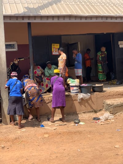 The scene is a street corner in Ondo, Nigeria, located at 7.251 latitude and 5.196 longitude. A group of people, mostly women, are standing in front of a building with a brown and white tiled roof. The building has a large doorway and a window. The people are gathered around a few buckets and plastic bags. One woman is wearing a purple dress and is standing next to a bucket. There are some scattered plastic bags and other debris on the ground. The people are standing on a concrete step, and the ground in front of the step is dirt. There are signs on the building, but they are not legible. There are several people standing near the entrance to the building. The people are all wearing clothes that are typical of the region. The weather appears sunny and warm.