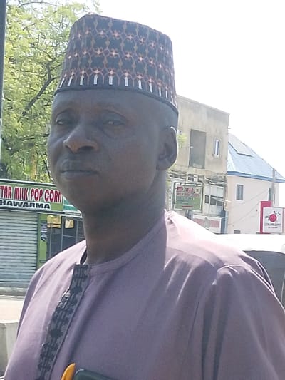 A dark-skinned adult male is depicted in a chest-up portrait against an urban backdrop in Gyadi Gyadi Arewa, Nigeria. He wears a patterned kufi cap featuring dark brown and light geometric designs, and a light purple traditional long-sleeved garment with a vertical black and grey patterned trim on the placket. A dark-colored object, possibly a mobile phone, and a bright yellow object are visible near his lower chest. His gaze is directed slightly to the viewer's left.
The background reveals a street scene under bright daylight. To the left, a shop with a closed metallic roll-down shutter displays a green sign reading "STAR MILK POP CORN SHAWARMA." Further left, green tree foliage is visible. To the right, a partially obscured red sign reads "RED STAR MILK POP CORN." Behind these shops, a multi-story, unfinished light brown concrete building with a single rectangular window is present, alongside another building with a dark-colored sloped roof. On the far right, a red and white sign with a circular logo is partially visible, along with a portion of a white vehicle.