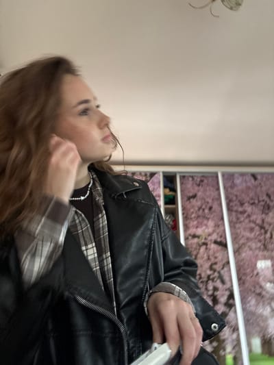 A young woman with long, wavy brown hair is shown in profile, looking to the left. She is wearing a black leather jacket over a plaid shirt and a dark top. A pearl necklace with a star pendant is visible around her neck. Her right hand is near her face, seemingly adjusting her hair or an earring. She is holding a white object, possibly a phone, in her left hand. The background appears to be an indoor setting with a closet or wardrobe behind her, decorated with a pink floral pattern, possibly cherry blossoms. The ceiling is visible at the top of the frame, and a light fixture is partially visible. The scene is likely taking place in Shepetivka, Ukraine.