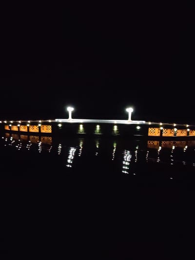 The image shows a bridge at night in Karmanovo, Russia. The bridge has orange sides with decorative cutouts, white lights along its span, and two taller lights in the center. Reflections of the bridge and lights are visible in the water below. The surrounding environment is dark, with minimal background detail visible.
