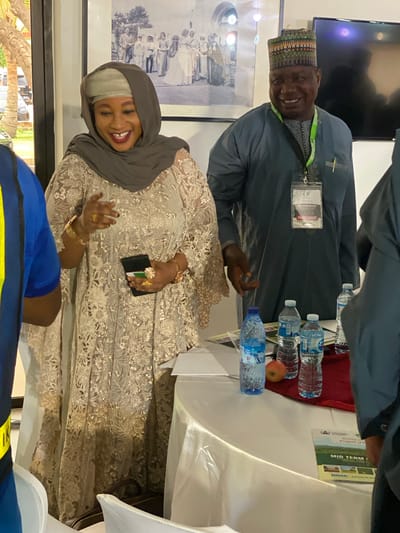 Two individuals are present in an indoor setting in what appears to be Kwarau, Nigeria. One is a woman wearing a hijab and an ornate, lace-like garment. She is smiling. The other person is a man wearing a traditional hat and a garment, with a name tag visible on his chest. They are positioned around a table covered with a white tablecloth, on which several bottles of water and a red apple are placed. A booklet or program with the words "MID TERM" on its cover is also on the table. In the background is a black-and-white framed photo.