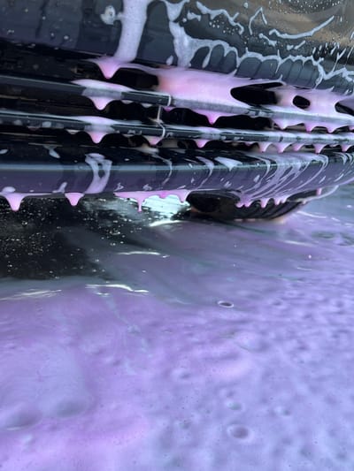 In Velyki Hai, Ukraine, a close-up, low-angle shot captures the underside of a dark-colored vehicle, likely a car, being washed. Pink foam and suds cascade down from the vehicle's grille and bumper, pooling on the ground beneath. The foam creates a frothy, iridescent layer that obscures the tire and the pavement. The scene suggests a car wash or a DIY car cleaning in progress, with the focus on the textures and colors of the soap and water.