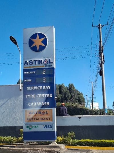 The image shows a large sign for an Astral petrol station in Nduma, Kenya, located at latitude -1.172, longitude 36.641. The sign is blue and white with a large yellow star, and it advertises unleaded petrol, diesel, service bay, tyre centre, car wash, Astral Mart, and a restaurant. The sign displays prices for unleaded petrol at 2 and diesel at 3, but does not specify the currency. A man in an orange hat and black shirt is sitting on a white wall behind the sign. The environment is urban, with power lines running across the sky and trees in the background.