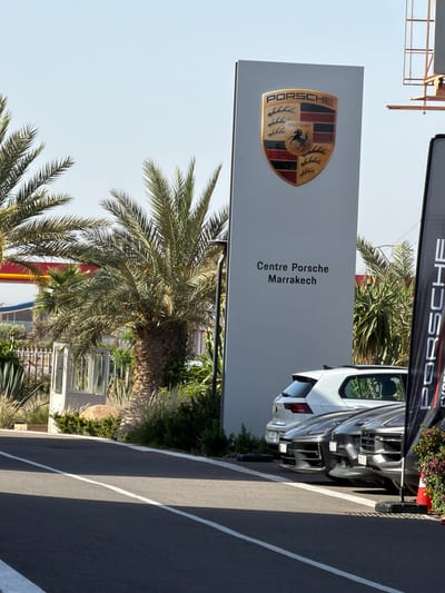 A sideways view of the Porsche Centre Marrakech, located in Ouahat Sidi Brahim, Morocco. A Porsche sign with the Porsche logo and "Centre Porsche Marrakech" text is prominently displayed. Palm trees surround the sign. A white Porsche car is parked nearby. A road with white lane markings runs alongside the scene.