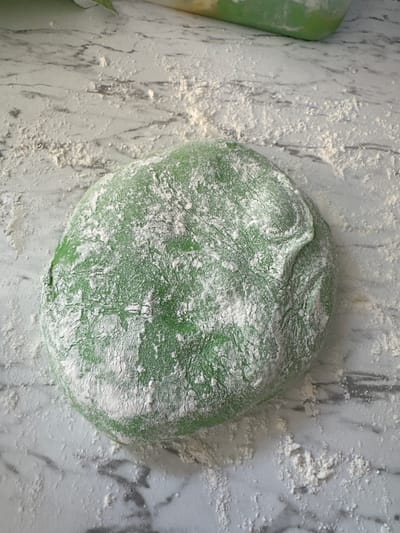 A green dough ball, dusted with white powder, rests on a marble countertop in Moortown, Ireland. The dough appears to be slightly flattened in the center, with visible creases and folds on its surface. Scattered around the dough are small piles and streaks of the same white powder, suggesting it is flour. The marble countertop exhibits a light gray veined pattern. The lighting in the image is diffused, casting soft shadows. There are no people or other living beings visible in the scene.