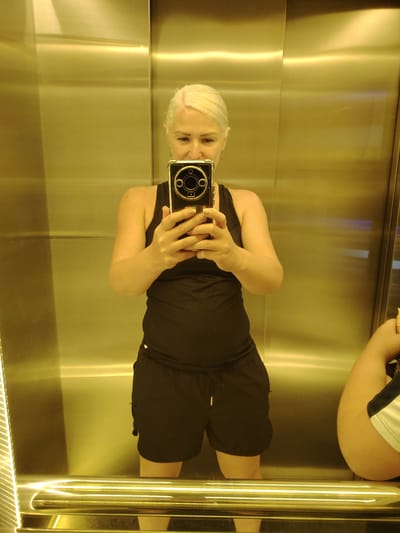 A woman with short blonde hair is taking a selfie in a golden elevator. She is wearing a black tank top and black shorts, holding a smartphone in her hands. Her face is visible, with a slight smile. The elevator walls are reflective and metallic, with vertical illuminated panels on the left side. Another person's arm and shoulder are visible in the reflection on the right side of the elevator, suggesting they are also present in the elevator. The floor of the elevator has a golden railing. The location is the city of Chaiky, Ukraine.