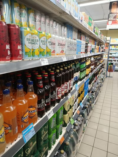 The image shows shelves of beverages in a supermarket in Khorol, Ukraine. The shelves are filled with various canned and bottled drinks, including beer, soda, and other beverages. The drinks are arranged in rows and are mostly colorful, with labels and branding visible. Some of the labels are in Ukrainian. The supermarket appears to be well-stocked and organized. The lighting is bright, typical of a retail environment, with fluorescent lights overhead. The floor is tiled with a light-colored ceramic tile. In the background, other shelves and aisles of the supermarket are visible, along with some shopping carts. There are no people prominently featured in the image, suggesting a focus on the products for sale.
