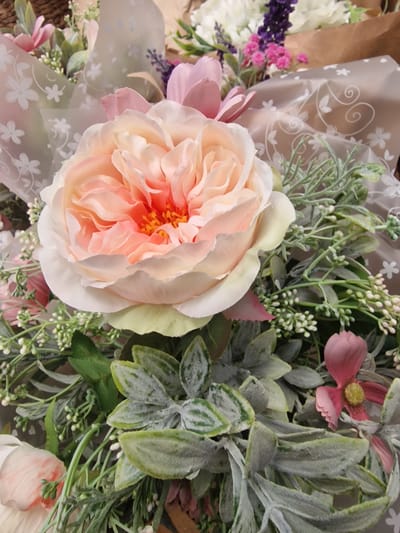 This is a close-up, overhead shot of a vibrant artificial floral arrangement. The primary subject is a large, intricately layered peach and cream rose positioned centrally, with its delicate petals unfurling outwards. Surrounding the rose are various other artificial flowers and foliage. To the left, a cluster of small purple flowers, possibly lavender, and bright pink blossoms are visible, interspersed with white blooms. To the right and behind the rose, a variety of greenery is present, including textured, dusty-looking leaves and wispy sprigs with tiny white buds.
The flowers are partially wrapped in a sheer, translucent material adorned with a delicate pattern of white floral designs and swirling lines. This wrapping adds a soft, decorative element to the arrangement. The overall impression is one of abundant, artificial beauty, likely intended for display or as a gift. There is no indication of people, specific weather conditions, time of day, or a clear outdoor location, as the focus is entirely on the static arrangement. The background is a mixture of brown material, likely part of the bouquet's packaging, and the soft tones of the floral elements. No visible text is present in the image.