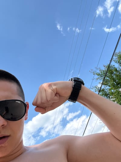 The image is a low-angle outdoor shot, primarily featuring a partially visible person against a blue sky. The person, who has short dark hair, is seen from the nose down, wearing dark Carrera polarized sunglasses. Their bare left shoulder and chest are visible, and their left arm is strongly flexed, showcasing a tensed bicep. A black, robust digital watch is worn on their left wrist. The background consists of a clear blue sky with scattered white cumulus clouds. Multiple parallel utility lines diagonally traverse the upper half of the frame from left to right. On the far right, a small portion of green foliage with reddish-brown branches is visible. There are no identifiable landmarks or signs to determine a specific city or country.