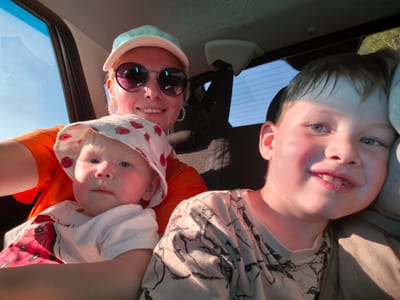 This is a medium shot, outdoor selfie taken from inside a vehicle. The sun is shining, creating a warm glow and strong shadows. In the foreground, two children are visible. The child on the right, a boy, is looking towards the camera with a slight smile. He has fair hair and is wearing a patterned t-shirt. To the left of the boy, a baby is nestled in the arms of a woman, looking to the side. The baby wears a white hat with a strawberry pattern and a white t-shirt with a red strawberry design. The woman, presumably their mother, is partially visible in the background, wearing an orange shirt, sunglasses, and a light-colored cap. She is smiling and looking at the camera. The background shows the interior of a car, including seats and a window with a view of a bright blue sky and trees. The scene suggests a family outing on a sunny day, possibly traveling by car through a rural or suburban area, given the presence of trees outside the vehicle. No specific details related to Karaiganovo, Russia are present in the image.
