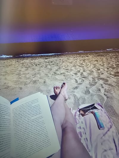The image shows a scene on a beach in Peníscola, Spain, as evidenced by the sandy beach and shoreline on the left side of the frame. A person is lying on a beach towel, visible with a purse and sunscreen bottle nearby. Their legs are visible, with their feet resting on the sand. The person appears to be reading a book.