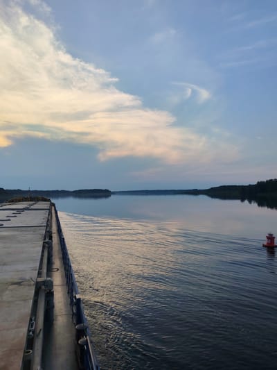 This is a wide-angle, eye-level, horizontal, outdoor, daytime photograph taken from the deck of a moving vessel. The scene captures a calm body of water, likely a river or lake, under a twilight sky. The water reflects the soft hues of the sky, with gentle ripples indicating the vessel's movement. On the left side of the frame, the edge of the vessel's deck is visible, characterized by weathered wooden planks and metal railings. The right side of the water features a distant tree-lined shore and a red buoy with the number "68" on it. The sky is a gradient of blue and pale yellow, with wispy clouds scattered across it, suggesting the setting or rising sun. There are no people visible in the image. The overall mood is serene and tranquil.