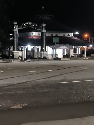 The image shows the entrance to a building at night, likely a mosque or religious center, in Sigli, Indonesia. A prominent sign above the entrance reads "39 PULO PISANG DUSUN BLANG LUMPAT KEC. JURIE." This indicates the location as Dusun Blang Lumpat, Pulo Pisang, in the Jurie sub-district of Sigli. The building has a distinctive architectural style with a red tiled roof and arched window-like features. White gates and pillars mark the entrance, with some decorative elements visible. A street with white lane markings runs in front of the building. A lamppost with an orange light is visible to the right of the entrance. The scene is illuminated by artificial lights, creating shadows and highlighting the building's features against the dark night sky. There are no people visible in the frame.