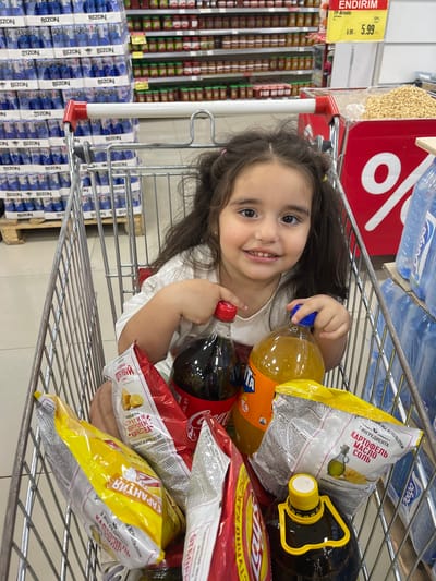 In this close-up, vertically oriented shot, a young girl with dark hair and bright eyes is lying down in a shopping cart filled with groceries. She is smiling broadly at the camera, her head propped up by her arm. The shopping cart contains various food items, including bags of chips, bottles of soda and juice, and a large bottle of what appears to be cooking oil. The background shows shelves stocked with more products, some in blue and white packaging, and a red display with a large percentage sign. The overall scene suggests a casual shopping trip. Visible text on product packaging includes brands like "BIZON," "Lay's," and "Fanta," along with Russian words that appear to describe the contents or ingredients of the snacks. There are also price tags visible, one indicating "5.99." The location context provided places this scene in Novxanı, Azerbaijan. The lighting suggests an indoor environment, likely a supermarket.